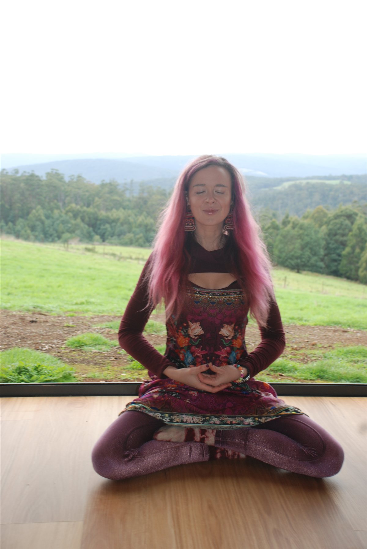 Melissa Warner seated cross-legged in meditation before a window of green hills