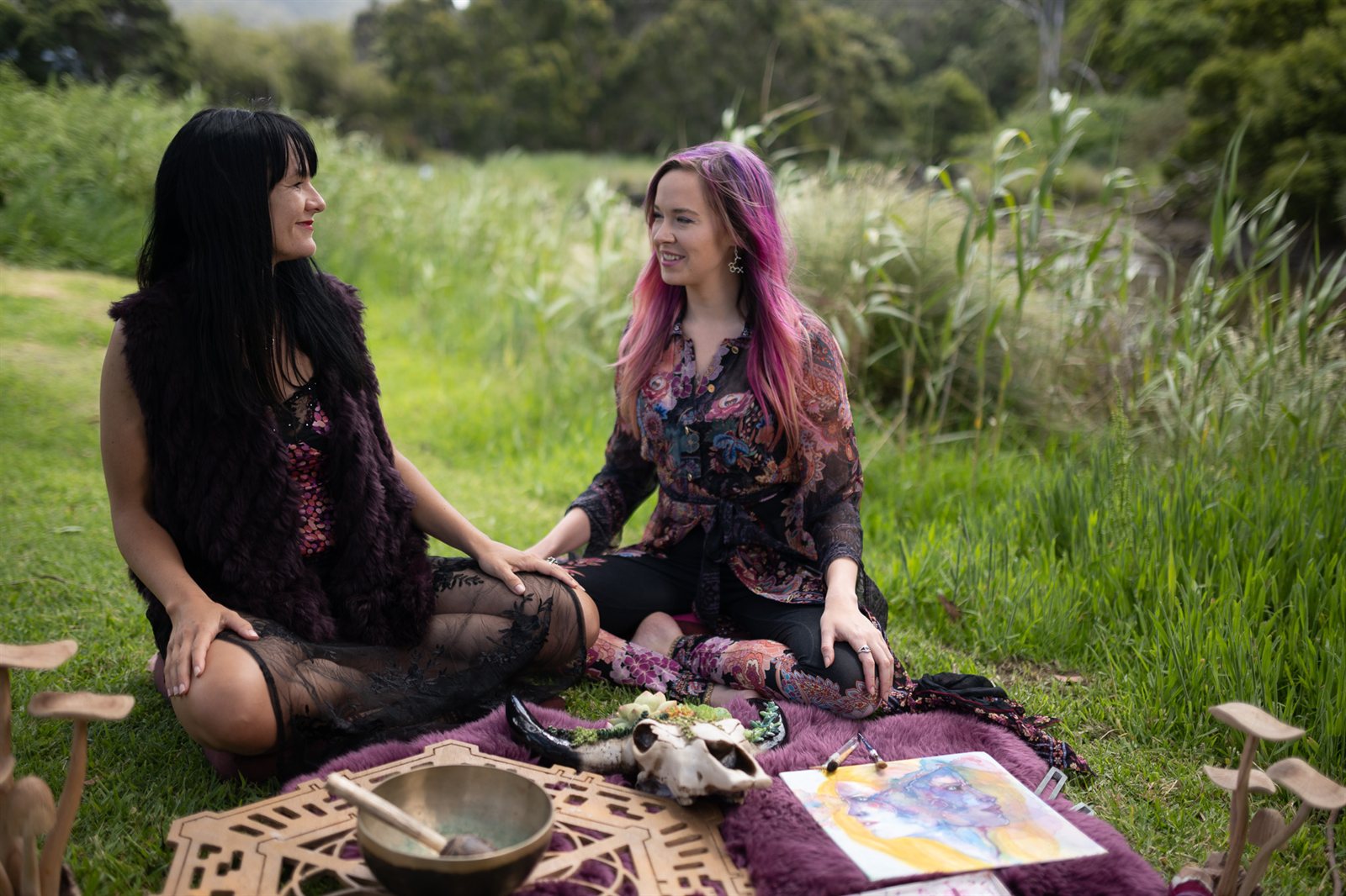 Anam Cara Centre co-founders seated together on a picnic rug in a green riverside meadow — Dr Alana (Lani) Roy with long black hair on the left, turned toward Melissa Warner with pink hair on the right, a brass singing bowl and carved wooden mushrooms before them